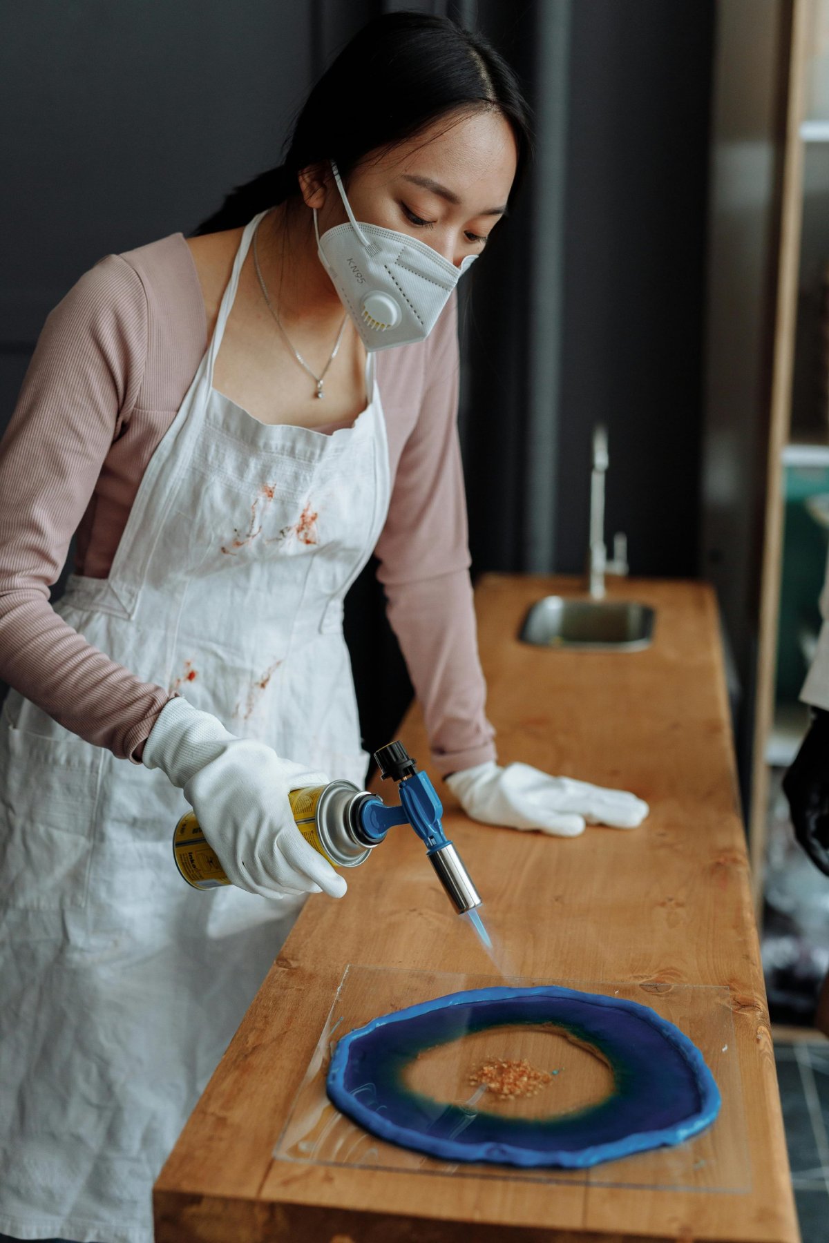 An artist uses a blowtorch to create intricate resin art on a wooden table, wearing protective gear indoors.