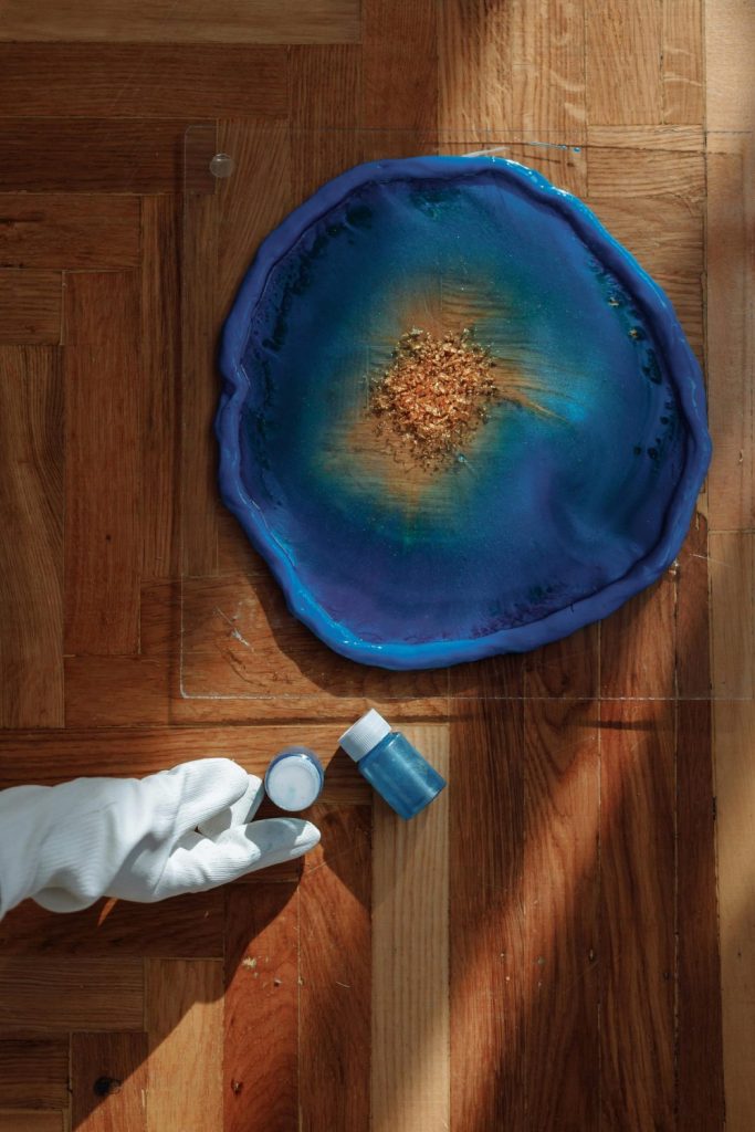 Colorful resin artwork displayed with artist holding pigment jar on wooden floor.